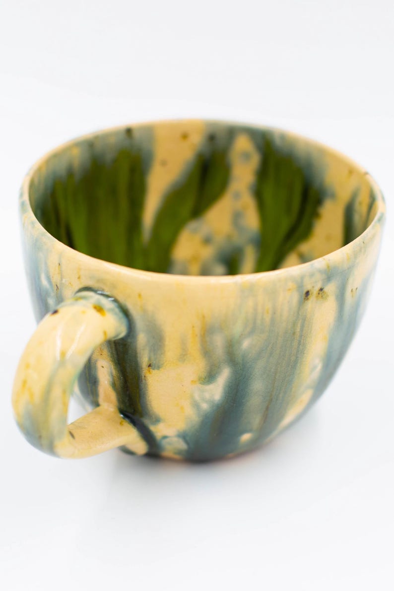 May include: Handmade ceramic mug with a unique glaze. The interior is a vibrant green, while the exterior features a blend of yellow, blue, and green hues. The mug has a rounded shape with a sturdy handle, perfect for enjoying your favorite beverage.