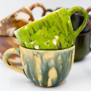 May include: Stacked ceramic mugs with unique, colorful glazes. The top mug is a vibrant green with white and black accents, resting on a mug with a yellow and blue glaze. Other mugs in the background have brown and dark green glazes.