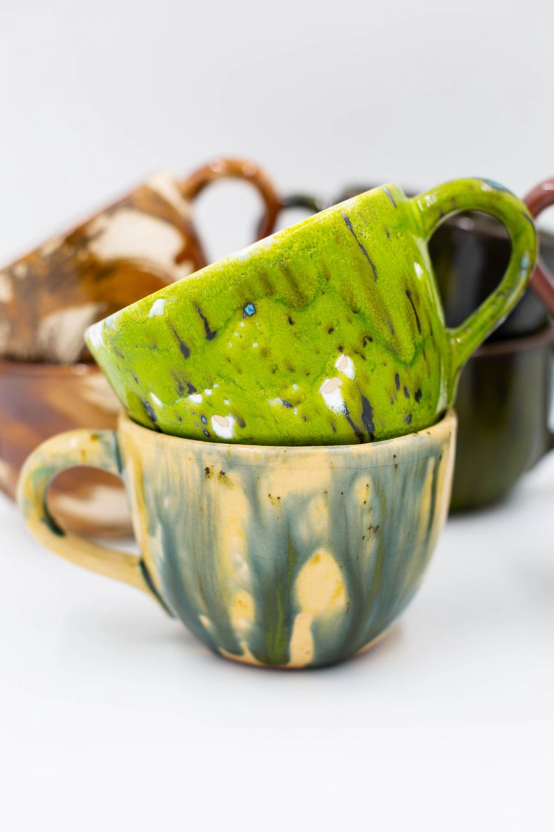 May include: Stacked ceramic mugs with unique, colorful glazes. The top mug is a vibrant green with white and black accents, resting on a mug with a yellow and blue glaze. Other mugs in the background have brown and dark green glazes.