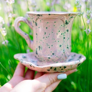 Puede incluir: Un juego de taza y platillo de cerámica rosa con un diseño moteado. La taza tiene un asa curvada y se apoya en un platillo a juego. El juego se sostiene en una mano sobre un fondo borroso de follaje verde y morado.