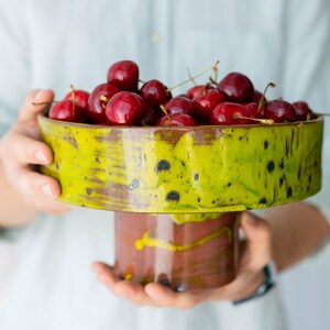May include: A ceramic pedestal bowl filled with ripe, red cherries. The bowl has a vibrant green and brown glaze with a unique, textured design. The pedestal base is a complementary brown colour.