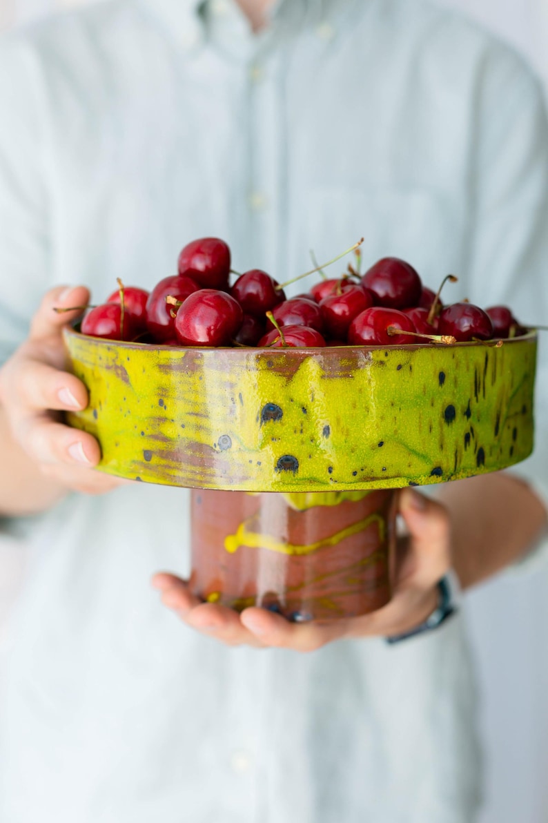 May include: A ceramic pedestal bowl filled with ripe, red cherries. The bowl has a vibrant green and brown glaze with a unique, textured design. The pedestal base is a complementary brown colour.