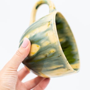 May include: Handmade ceramic mug with a unique glaze. The mug features a blend of yellow, green, and blue hues, creating a marbled effect. The handle is a matching color. A perfect addition to any kitchen or coffee collection.