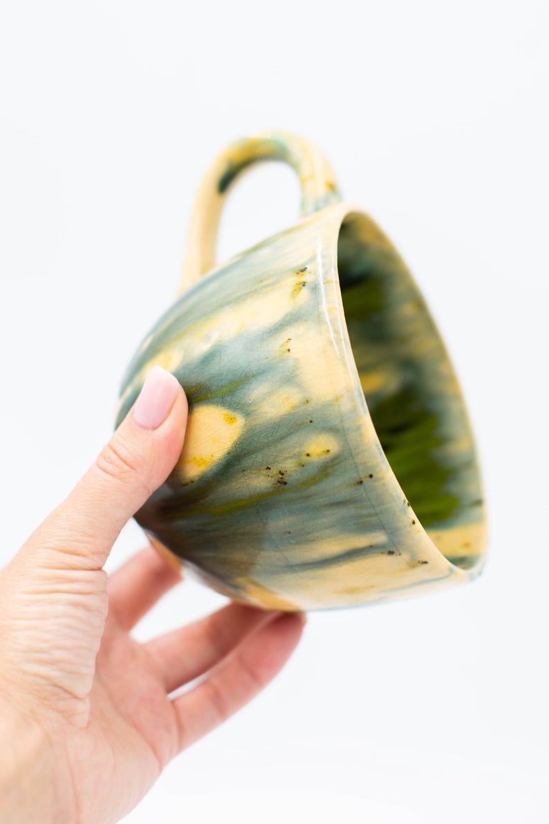 May include: Handmade ceramic mug with a unique glaze. The mug features a blend of yellow, green, and blue hues, creating a marbled effect. The handle is a matching color. A perfect addition to any kitchen or coffee collection.