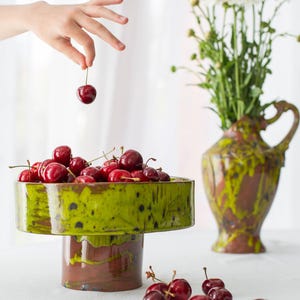 May include: A still life composition with a green and brown ceramic pedestal bowl filled with red cherries. A hand holds a cherry above the bowl. A vase of white flowers is in the background. Cherries are scattered on the table.
