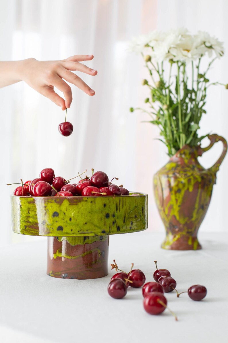 May include: A still life composition with a green and brown ceramic pedestal bowl filled with red cherries. A hand holds a cherry above the bowl. A vase of white flowers is in the background. Cherries are scattered on the table.