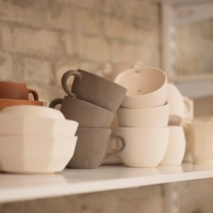 May include: A collection of ceramic cups, bowls, and teacups in various shades of brown, gray, and white, arranged on white shelves. The cups have handles, and the bowls have geometric shapes. A green bowl is visible in the background.