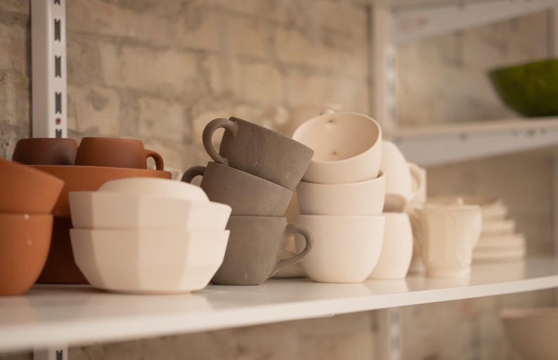 May include: A collection of ceramic cups, bowls, and teacups in various shades of brown, gray, and white, arranged on white shelves. The cups have handles, and the bowls have geometric shapes. A green bowl is visible in the background.