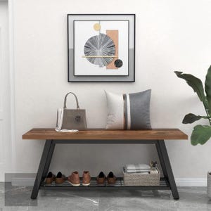 Puede incluir: Un banco de madera con estructura de metal negro, con una balda inferior para zapatos y una cesta de mimbre. Un bolso gris y un cojín decorativo están sobre el banco. Una obra de arte abstracta enmarcada cuelga encima del banco.