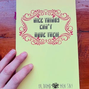May include: A yellow notebook with the text "NICE THINGS CAN'T HAVE THEM" in a decorative pink frame. Below the text is the phrase "OR. DUMB MEN SAY" in black. A hand is visible on the left side of the notebook.