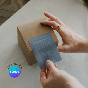 Könnte beinhalten: Eine braune Pappschachtel mit einem blauen Etikett mit der Aufschrift "YOUR BRAND NAME" und "SEA SALT & ORCHID". Das Etikett wird von den Händen einer Person gehalten. Das Canva-Logo befindet sich unten links.
