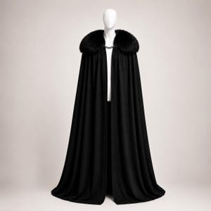 May include: A long, black cloak with a faux fur collar and a chain closure. The cloak is displayed on a mannequin against a neutral background. The fur collar is dark and plush, adding a touch of luxury to the garment.