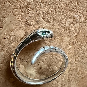 May include: A silver snake ring featuring a green gemstone eye. The ring is designed to coil around the finger, with the snake's head and tail forming the ends. The ring has a textured, scale-like pattern.
