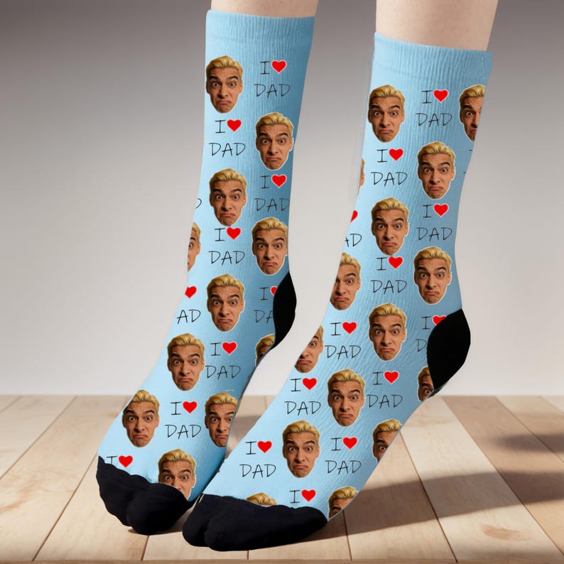 Może przedstawiać: Jasnoniebieskie skarpetki z powtarzającym się wzorem twarzy, napisami "I ❤️ DAD" i czerwonymi sercami. Skarpetki mają czarne pięty i palce, prezentowane na drewnianej powierzchni.