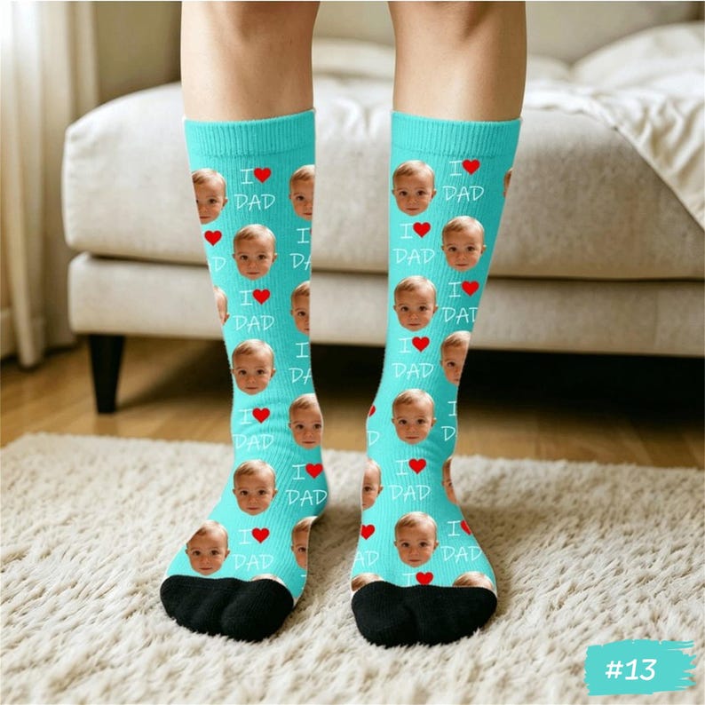 Może przedstawiać: Para turkusowych skarpet z powtarzającym się wzorem twarzy dziecka, czerwonych serc i napisem "I ❤️ DAD". Skarpety mają czarne pięty i palce. Skarpety są na jasnej wykładzinie.
