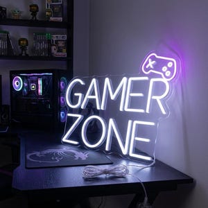 Könnte beinhalten: Ein weißes Neon-Schild "GAMER ZONE" mit einem Controller-Symbol, das in Lila leuchtet. Das Schild steht auf einem schwarzen Schreibtisch mit einem Gaming-Mauspad. Ein Computerturm und Regale mit Figuren und Spielen sind im Hintergrund.