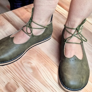 May include: Olive green leather shoes with a lace-up design. The shoes have a rounded toe and a decorative cutout on the side. The soles are cream-coloured with a black and white trim. The shoes are on a wooden surface.