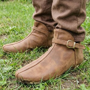 Puede incluir: Botines de cuero marrón con cierre de hebilla. Las botas tienen punta afilada y se usan con pantalones marrones. Las botas están sobre una superficie cubierta de hierba.