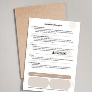 Puede incluir: Una tarjeta blanca con instrucciones de seguridad alemanas, titulada "Sicherheitshinweise", se muestra en un sobre marrón. La tarjeta detalla las precauciones de seguridad para varios artículos, incluyendo velas, madera y flores secas.