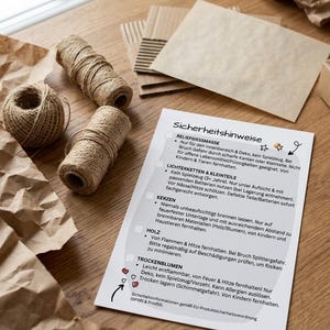 Pode incluir: Uma natureza morta com materiais de artesanato sobre uma superfície de madeira. A imagem inclui rolos de barbante, papel castanho amassado, pedaços de cartão e um cartão branco com texto alemão sobre precauções de segurança.