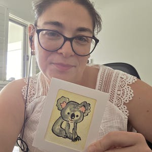 May include: A small rectangular white card featuring a hand-drawn illustration of a grey koala with pink inner ears and a black nose, set against a pale yellow background, held up by a person wearing a white sleeveless top with lace trim.