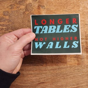 May include: A rectangular sticker with the text "LONGER TABLES NOT HIGHER WALLS" in red and light blue letters on a dark green background. The sticker is held by a hand against a wooden surface.