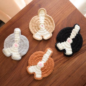 May include: Four handmade crochet coasters in the shape of dog butts. The coasters are in various colors: gray, tan, brown, and black, each with a white stripe and a pink accent. They are arranged on a wooden surface.