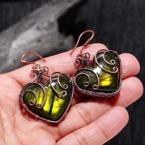 May include: A pair of heart-shaped earrings. Each earring features a green gemstone with yellow highlights, encased in copper wire. The wire is intricately woven around the stone and forms decorative swirls at the top. The earrings have copper ear wires.