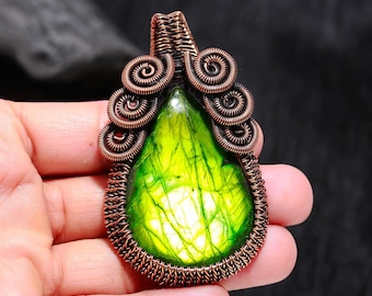 May include: A handcrafted copper wire pendant featuring a vibrant, pear-shaped green gemstone. The gemstone is encased in intricate wirework, with decorative spiral accents at the top. The pendant is held in a hand, showcasing the craftsmanship and unique design.