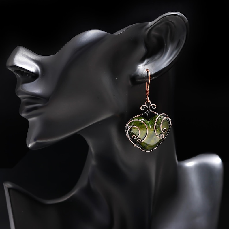 May include: A handmade earring featuring a heart-shaped green stone encased in a copper wire frame. The earring hangs from a copper hook, with decorative swirls at the top of the heart. The background is black, highlighting the jewelry.
