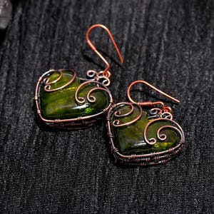 May include: A pair of handmade earrings featuring heart-shaped green glass stones encased in copper wire. The wirework forms a decorative frame around the glass, with intricate swirls and loops. The earrings have copper ear wires.