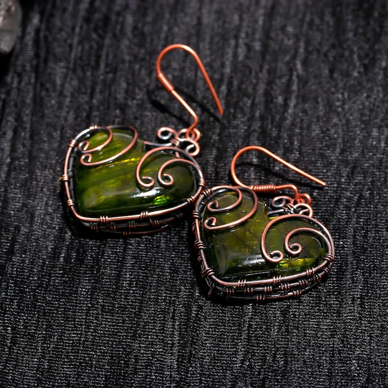 May include: A pair of handmade earrings featuring heart-shaped green glass stones encased in copper wire. The wirework forms a decorative frame around the glass, with intricate swirls and loops. The earrings have copper ear wires.