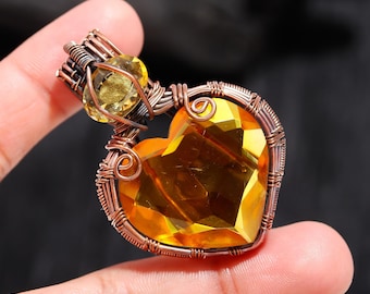 May include: A heart-shaped pendant with a large, faceted yellow gemstone. The gemstone is encased in copper wire, with a smaller yellow stone at the top. The pendant is held by a person's fingers.