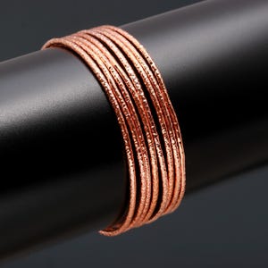 May include: A collection of copper-toned bangles with a textured finish. The bangles are presented on a black cylindrical stand against a dark grey backdrop. The jewellery has a hammered appearance.
