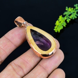 May include: A teardrop-shaped pendant with a deep purple gemstone set in a gold-colored frame. The pendant has a copper-colored bail and is held against a dark background. The gemstone is a rich, saturated purple color.