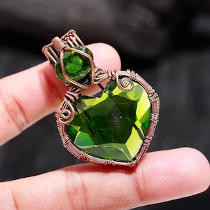 May include: A handcrafted pendant featuring a heart-shaped, faceted green gemstone. The gemstone is encased in copper wire, with a smaller green stone above. The wirework includes decorative swirls and a bail for a chain.