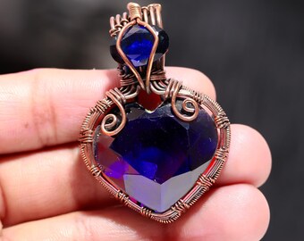 May include: A heart-shaped pendant with a large, faceted, deep blue gemstone. The gemstone is encased in copper-colored wire, with intricate swirls and loops. A smaller blue stone sits above the heart, also wire-wrapped. The pendant is held in a hand.