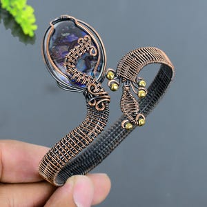 Pode incluir: Uma pulseira de punho artesanal em fio de cobre. A pulseira apresenta uma pedra oval iridescente emoldurada em um fio. O design inclui uma intrincada tecelagem de fios e pequenos detalhes dourados, criando uma peça de joalheria única e artística.