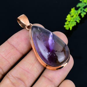 May include: A teardrop-shaped pendant featuring a polished, translucent purple gemstone with brown undertones, set in a rose gold-colored metal frame. The pendant has a loop at the top for a chain. The gemstone is smooth and has a glossy finish.