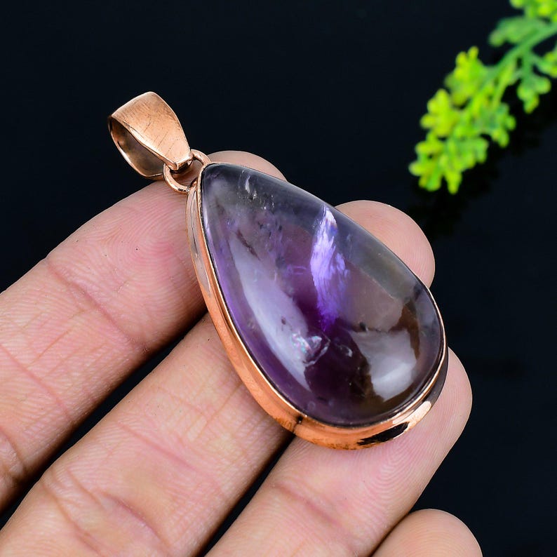 May include: A teardrop-shaped pendant featuring a polished, translucent purple gemstone with brown undertones, set in a rose gold-colored metal frame. The pendant has a loop at the top for a chain. The gemstone is smooth and has a glossy finish.