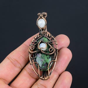 Puede incluir: Colgante artesanal con una piedra verde y roja, acentuado con dos piedras blancas. Las piedras están encapsuladas en alambre de cobre, creando un diseño artístico. El colgante mide aproximadamente 5 cm de largo.