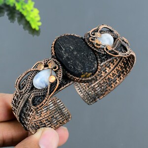 Op de afbeelding: Handgemaakte koperdraad manchetarmband met een grote, ovale zwarte steen en twee kleinere, ronde, lichtblauwe stenen. De armband heeft een open ontwerp met ingewikkeld draadwerk en een geweven textuur.