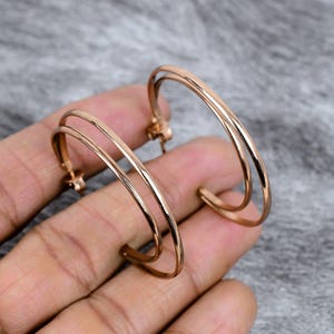 Puede incluir: Un par de pendientes de aro de oro rosa. Cada pendiente presenta un diseño de doble capa, con dos aros paralelos conectados en el poste. Los pendientes tienen un acabado pulido y un tono metálico cálido. La joyería se muestra sobre un fondo neutro.