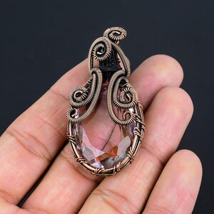 Puede incluir: Un colgante artesanal con una piedra preciosa facetada en forma de lágrima, en tonos rosas y morados. La piedra está engastada en intrincado alambre de cobre, con espirales decorativas y un acento de alambre negro en la parte superior. El colgante se sostiene en una mano.