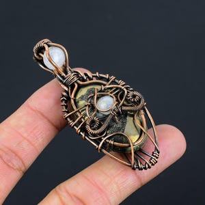 May include: A handcrafted copper wire pendant featuring a central stone with a mix of gold and dark grey hues, accented by two small, round, white stones. The pendant is intricately wired, showcasing a unique, artistic design. The overall aesthetic is earthy and bohemian.