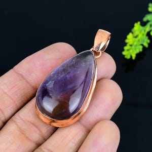 May include: A teardrop-shaped amethyst pendant with a copper-colored metal frame and bail. The polished purple gemstone has darker streaks and a reflective surface. The pendant is designed for use with a necklace.