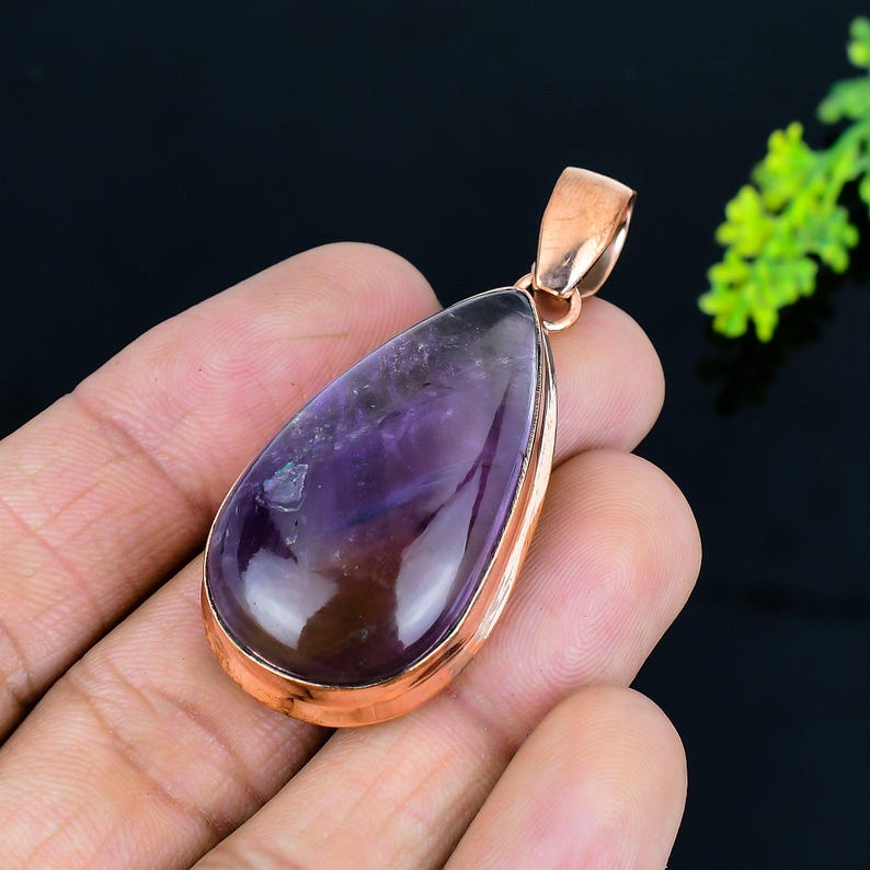 May include: A teardrop-shaped amethyst pendant with a copper-colored metal frame and bail. The polished purple gemstone has darker streaks and a reflective surface. The pendant is designed for use with a necklace.