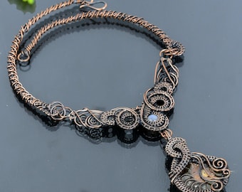 Puede incluir: Un collar artesanal de alambre de cobre con intrincados diseños en espiral y circulares. El collar presenta un colgante con una piedra fosilizada. El collar es una pieza de joyería única.