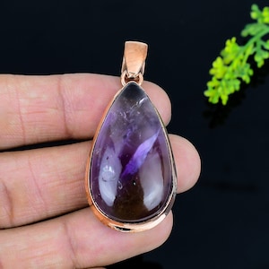 May include: A teardrop-shaped pendant with a polished, translucent purple gemstone. The stone is set in a copper-colored metal frame with a matching bail. The pendant is held against a dark background.