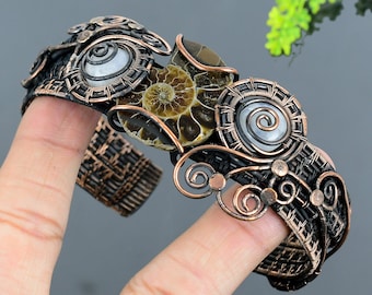 May include: A handcrafted copper wire bracelet featuring a fossilized ammonite and two spiral-shaped, pearlescent stones. The intricate wirework is in a dark bronze colour, with decorative swirls and a woven band.
